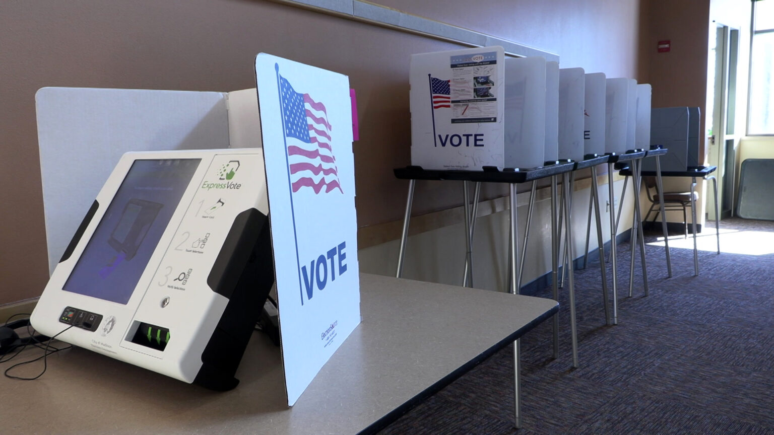 An electronic voting machine with a three-sided folding paper privacy screen with an illustration of a U.S. flag and the word VOTE site on a table next to a wall, with four more voting booths with collapsible metal and plastic tables topped with privacy screens along the wall in the background, in a room with short-pile carpet and is illuminated by sunlight from large plate-glass windows on the rear wall.