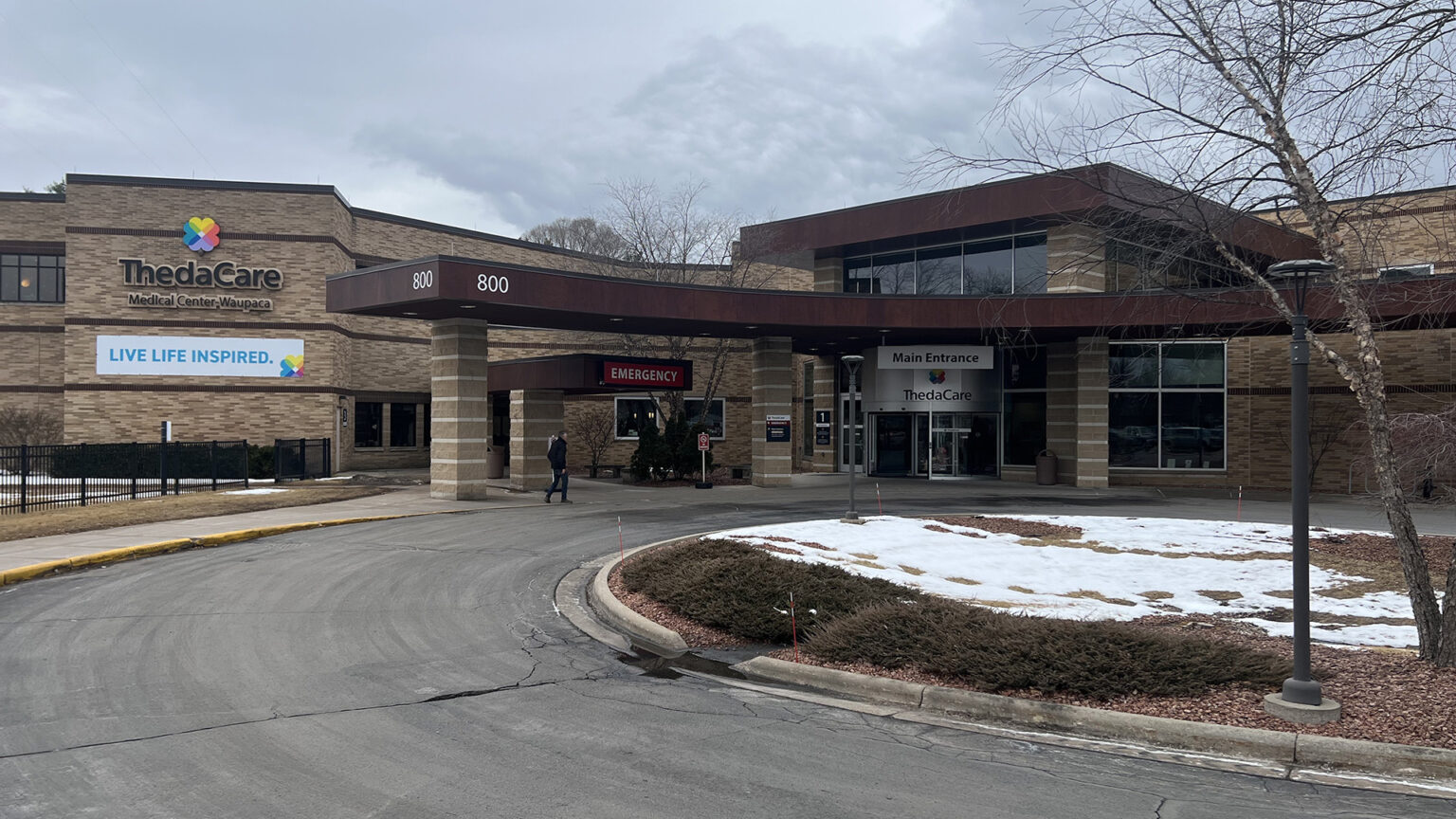 A person walks under an awning that extends over sidewalk along a curved driveway in front of a two-story brick building with multiple wings, entrances with signs reading Emergency and Main Entrance, and a banner reading Live Life Inspired, with a leafless tree, snow-covered lawn and landscaping plants in a median surrounded by the road, with more trees behind the building in the background under a cloudy sky.