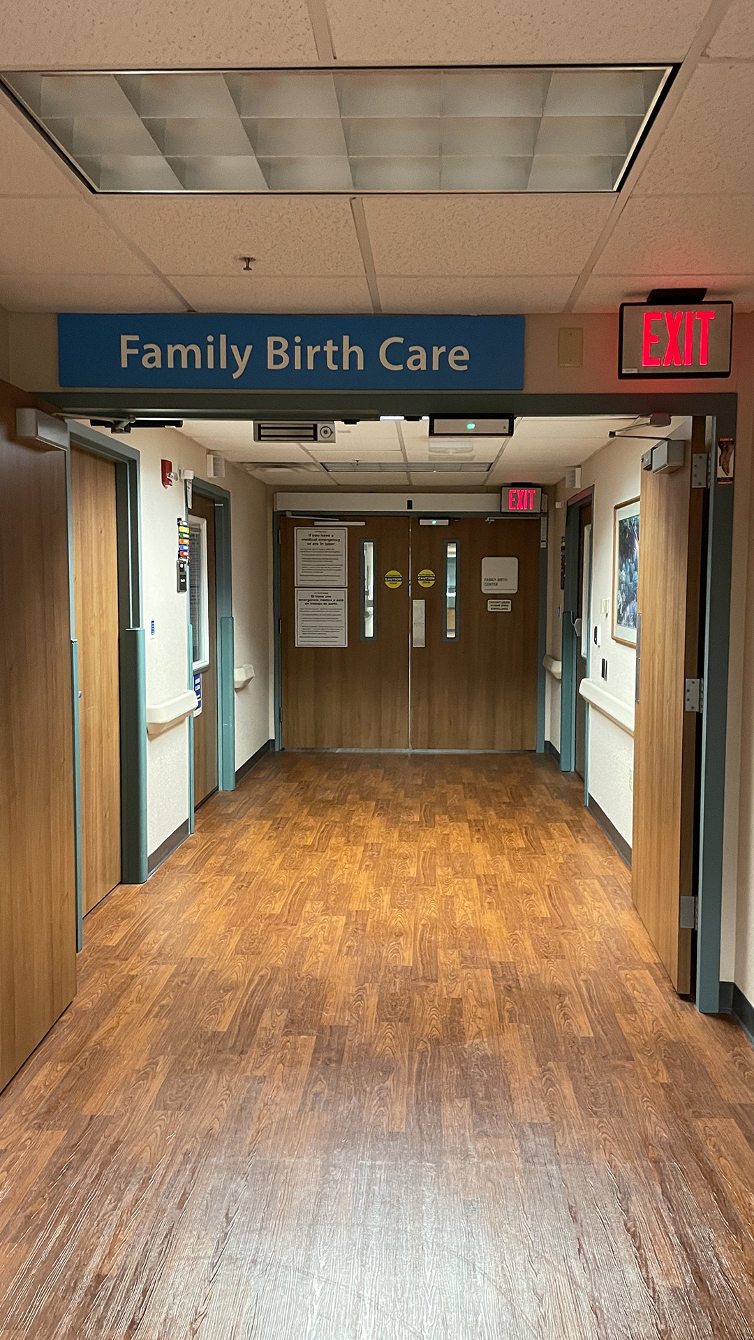 An illuminated EXIT sign and another sign reading "Family Birth Care" are visible above a doorway with open pneumatic double doors leading to another pair of closed pneumatic double doors accompanied by an illuminated EXIT sign," in a hallway with a wood floor and drop-panel ceiling.