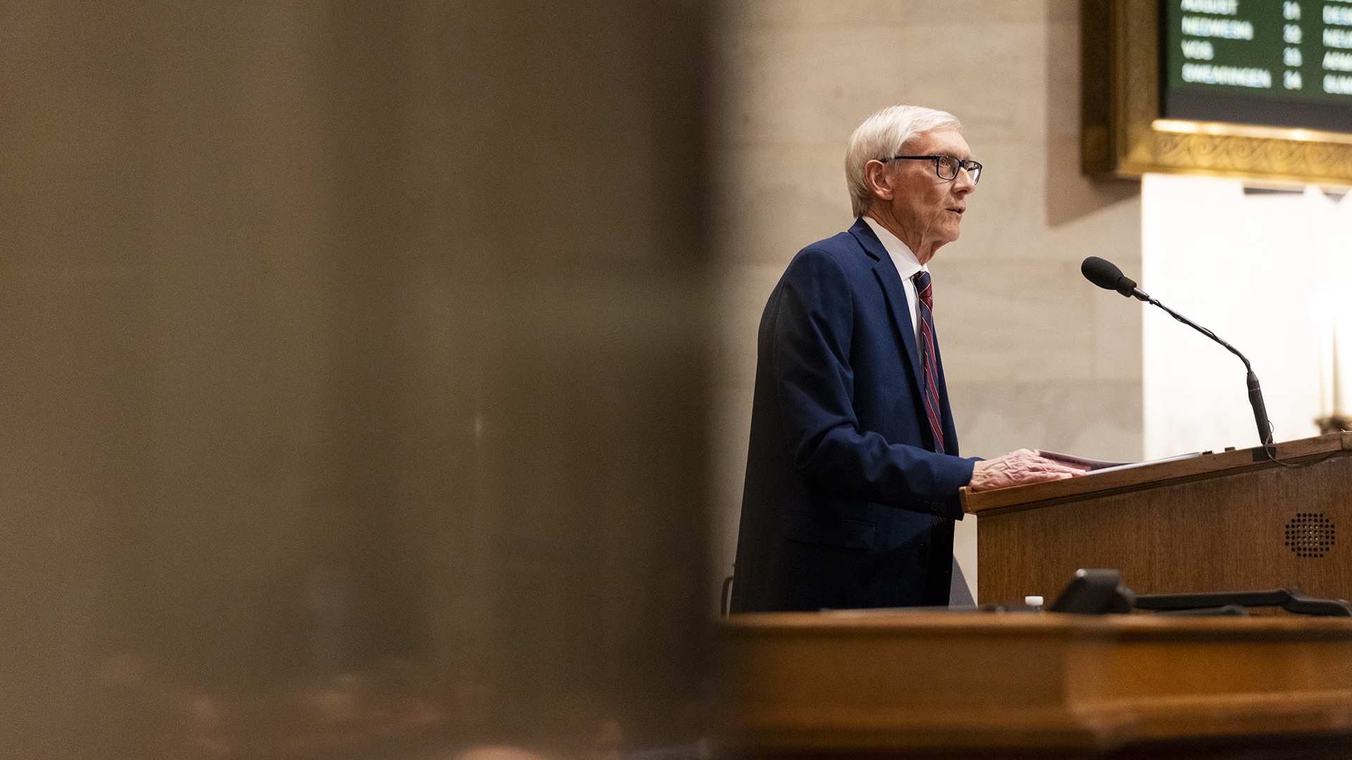 Tony Evers speaks into a microphone mounted to the top of a wood podium, while standing in a room with marble masonry and an out-of-focus digital vote register, with an out-of-focus polished metal surface in the foreground.