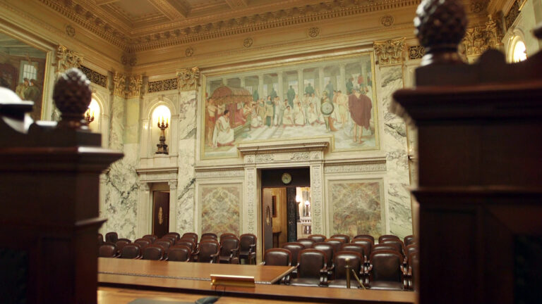 The backs of two out-of-focus high-backed wood chairs topped with carved pinecones stand facing the surface of a judicial dais, a wood table and multiple rows of leather-upholstered wood arm chairs, in a room with marble masonry, illuminated brass lighting fixtures in wall niches, multiple large paintings on the wall, and painted ceiling molding.