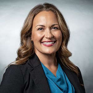 Smiling woman with wavy light brown hair wearing a black blazer and teal blouse against a gray gradient background.