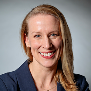 Smiling woman with long blonde hair wearing a navy blazer and gold necklace against a gray background.