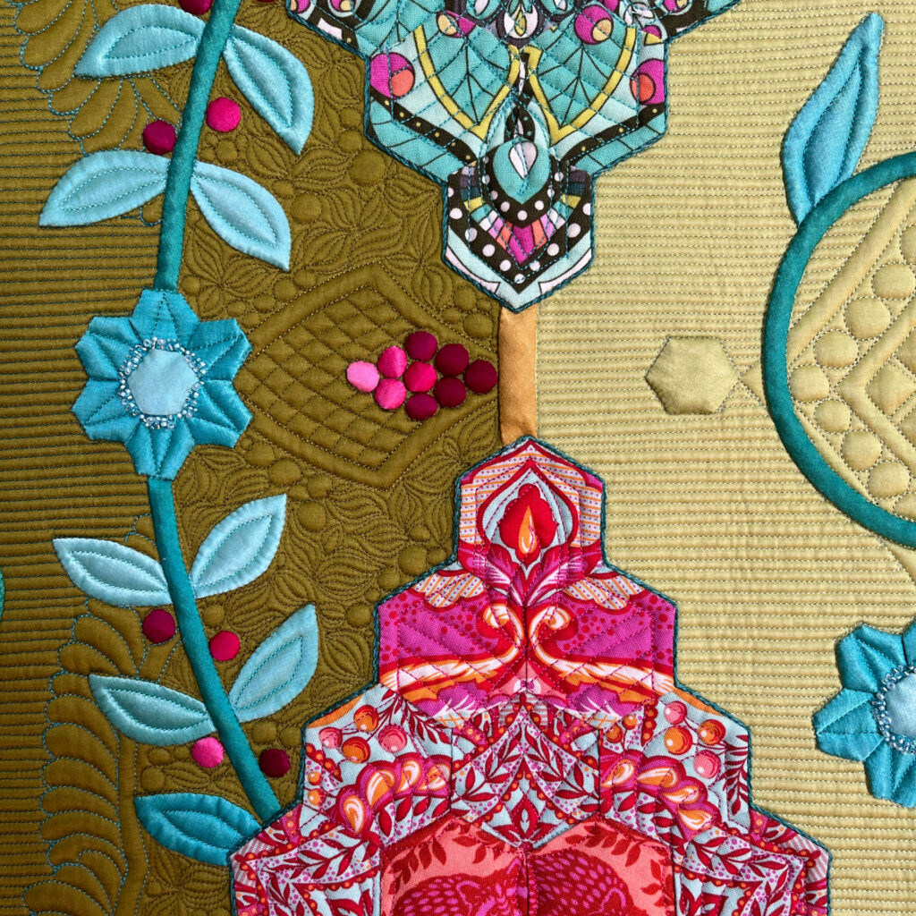 Close-up of a colorful quilt section featuring turquoise flowers, stylized vines, and an ornate pink-red motif framed by golden and green stitched textures.