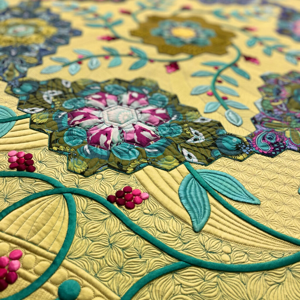 Intricate quilt detail with layered hexagons, floral patterns, and stitched leaves on a yellow background with teal vines and magenta berry-like accents.