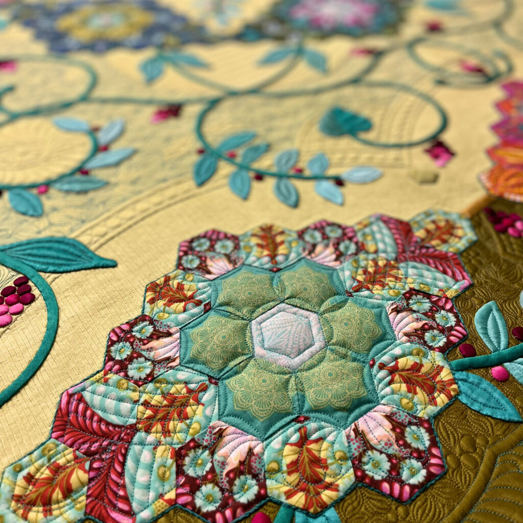 Quilted hexagon rosette with teal, pink, and red patterns surrounded by stitched leaves and vines on a yellow and olive background.