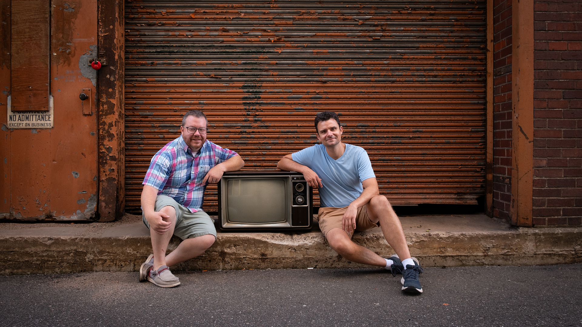 Two men sit on a sidewalk in front of a rusted garage door. Each of them rests their arm on a vintage television.