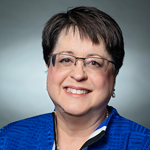 Smiling woman with short dark hair and glasses wearing a blue textured jacket and pearl earrings against a gray background.