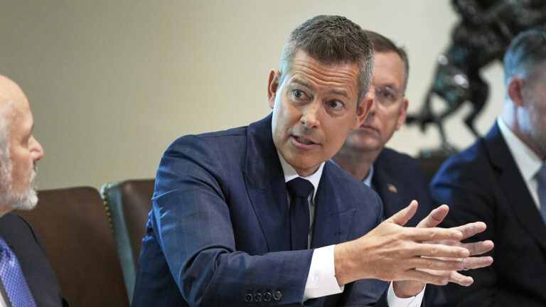 Sean Duffy gestures with both hands while turning to his right and speaking while seated in a high-backed leather chair, with other people sitting on either side of him in a room with an out-of-focus bronze statue in the background.
