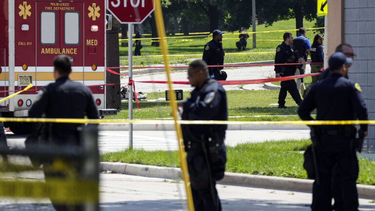 Multiple police officers stand in different streets near an intersection surrounded by multiple placements of crime scene tape, with an ambulance  parked near them, with multiple trees in a field in the background.
