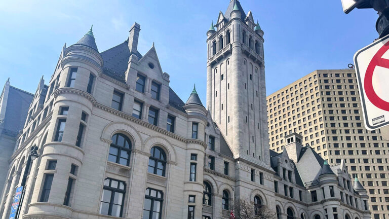 Sunlight illuminates and casts shadows on a multi-story Richardsonian Romanesque-style masonry building with multiple turrets, gables and a central tower, with other buildings behind it and a portion of a traffic sign in the foreground.