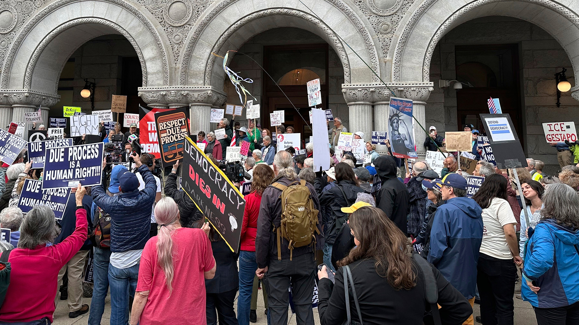 A group of people, with many holding printed and hand-written signs in their hands, stand in front of a stone masonry building with multiple Romanesque-style arches in front of doors.