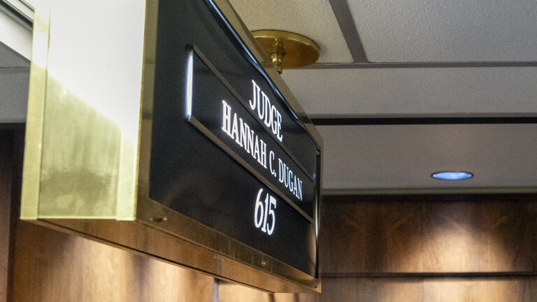 A ceiling-mounted brass enclosed sign with the words Judge Hannah C. Dugan and the number 615 stands outside the wood-paneled doors to another room.