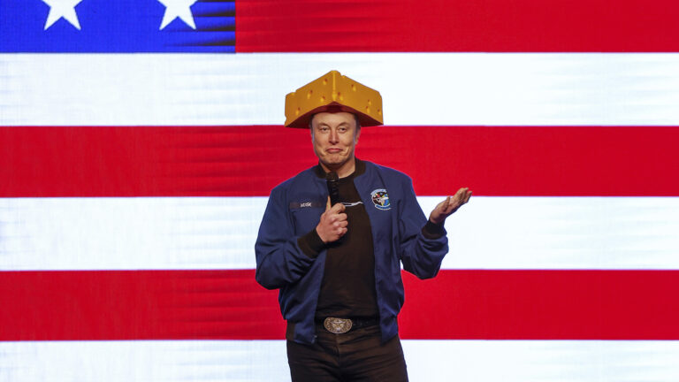 Elon Musk speaks with a wireless microphone in his right hand and gesturing with left hand, while hearing a Cheesehead hat and standing in front of a projection of the U.S. flag.