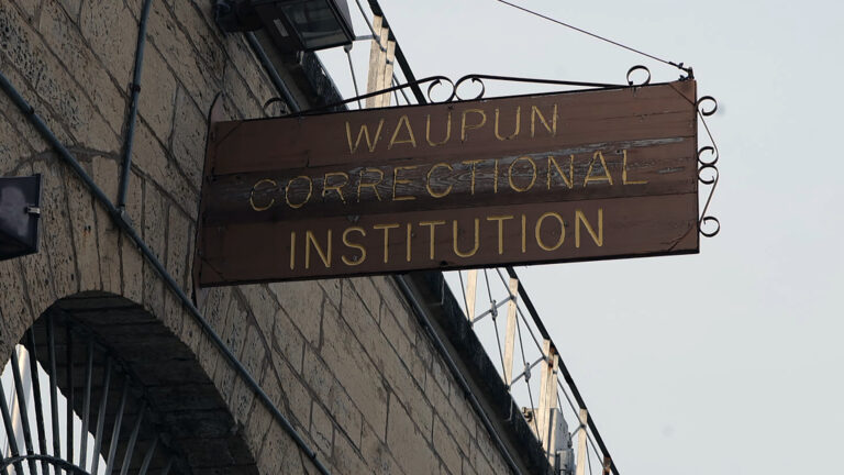 A wood sign with painted inlay words reading Waupun Correctional Institution is attached to the side of a masonry wall above  an arch filled with a sunburst metal grille.