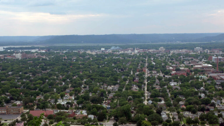 An aerial image shows a wooded cityscape with a grid street pattern and multiple neighborhoods of buildings along with a double-arched bridge over a river and adjacent bottoms, with bluffs on the horizon in the background. An aerial image shows a wooded cityscape with a grid street pattern and multiple neighborhoods of buildings along with a double-arched bridge over a river and adjacent bottoms, with bluffs on the horizon in the background.