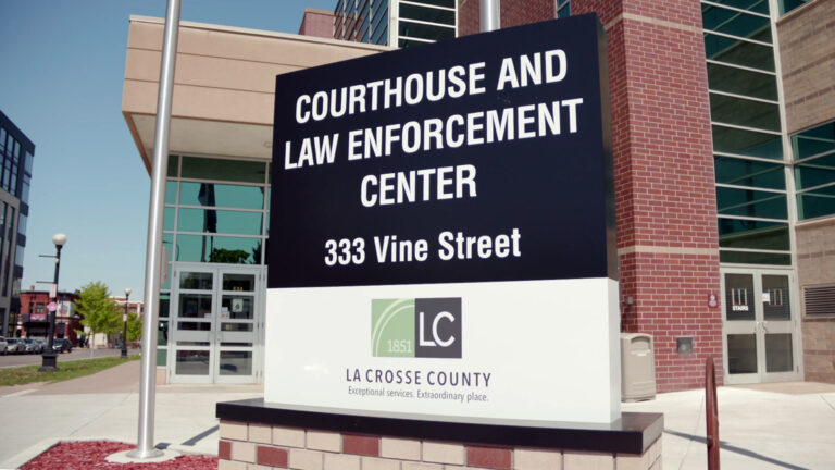 A sign with the words Courthouse and Law Enforcement Center, the address 333 Vine Street and a wordmark graphic above the words La Crosse County and Exceptional services. Extraordinary place. stands on top of a brick base next to a metal flagpole, in front of a multi-story brick-and-glass walled building, with other buildings and street lights to one side in the background.