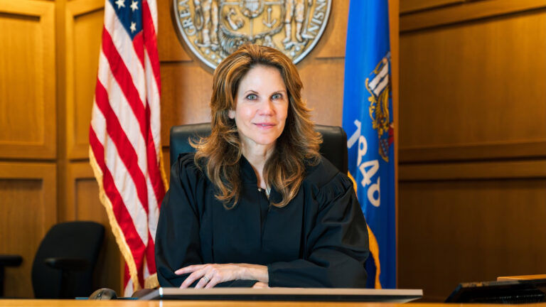 Chris Taylor sits in a high-backed leather chair on a judicial dais, with U.S. and Wisconsin flags behind her flanking a wall-mounted metal Great Seal of the State of Wisconsin, in a room with wood-paneled walls.