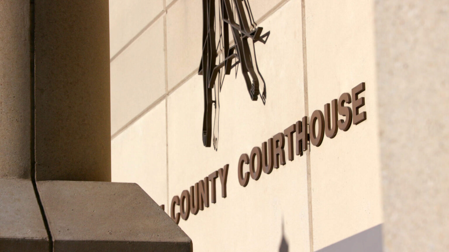 A sign that reads in part County Courthouse and the bottom of a stylized flat, wire sculpture are mounted on the masonry wall of a Brutalist-style building.