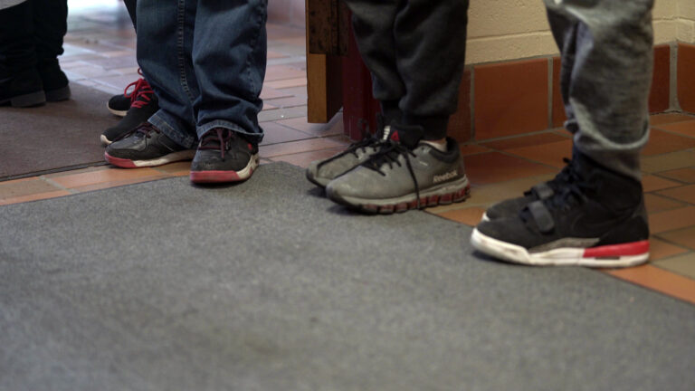 The shoes of multiple children are standing on rubber backed carpet maps in a hallway with a brick floor.