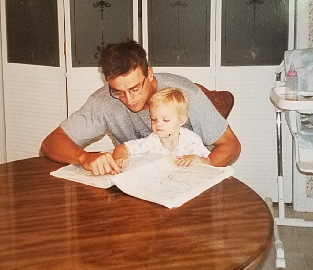 A baby sits on her father's lap. They're looking at an atlas.