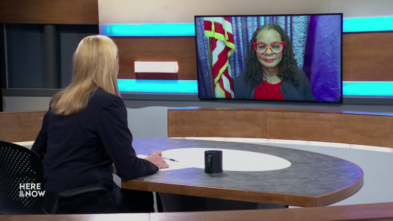 Frederica Freyberg sits at a desk on the Here & Now set and faces a video monitor showing an image of Gwen Moore.