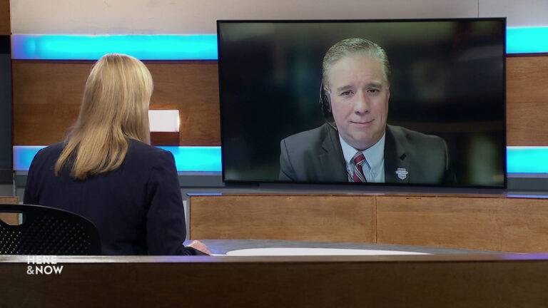 Frederica Freyberg sits at a desk on the Here & Now set and faces a video monitor showing an image of Darry Morin.
