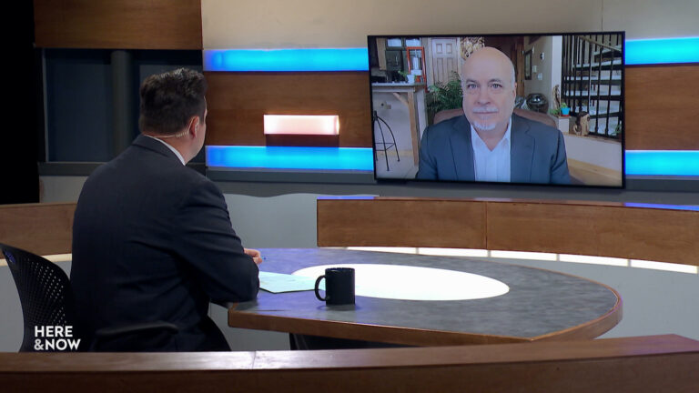 Frederica Freyberg sits at a desk on the Here & Now set and faces a video monitor showing an image of Mark Pocan.