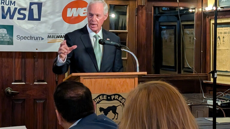 Ron Johnson gestures with his right hand and speaks into a microphone mounted to the top of a wood podium while standing in a room with a logo banner, a wood door, wood-framed windows, and a framed front page of a newspaper, with the backs of the heads of seated people in the foreground.
