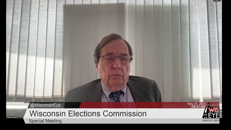 A video still image shows Bob Spindell sitting in an office with sunlight and shadows visible on closed vertical blinds in the background, with a video graphic at bottom including the text Wisconsin Elections Commission and Special Meeting.