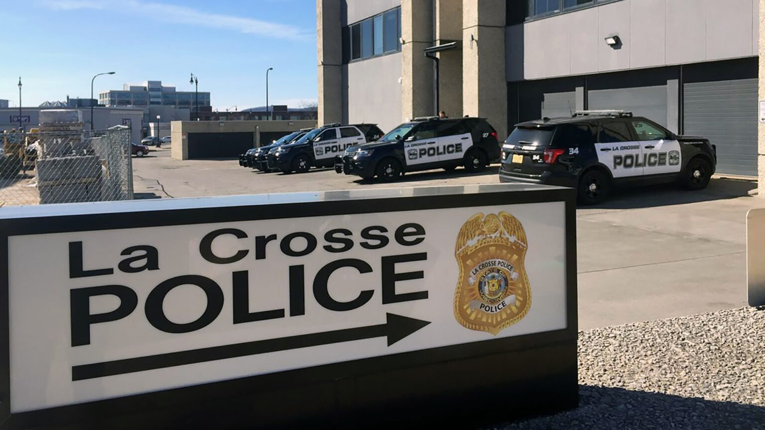 An unilluminated backlit sign with the words La Crosse Police and an arrow next to a police badge stands in front of a parking lot with multiple parked law enforcement vehicles next to a multi-story Brutalist-style building, light poles and other buildings in the background to one side. An unilluminated backlit sign with the words La Crosse Police and an arrow next to a police badge stands in front of a parking lot with multiple parked law enforcement vehicles next to a multi-story Brutalist-style building, light poles and other buildings in the background to one side.