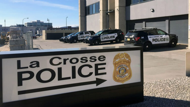 An unilluminated backlit sign with the words La Crosse Police and an arrow next to a police badge stands in front of a parking lot with multiple parked law enforcement vehicles next to a multi-story Brutalist-style building, light poles and other buildings in the background to one side.