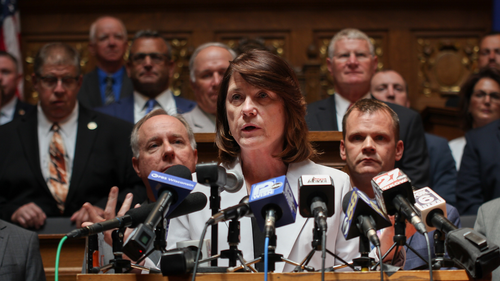 Mary Felzkowski speaks into multiple microphones with the flags of different media organizations mounted to the top of a wood podium, with Robin Vos, Devin LeMahieu and other people standing behind her on multiple levels of a legislative dais, in a room with a U.S. flag and carved wood-paneled walls.
