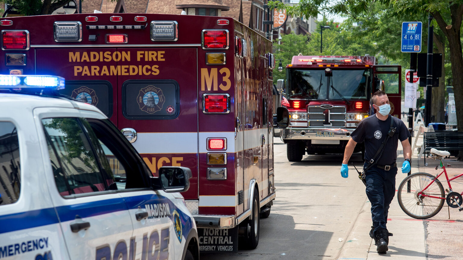 A first responder wearing nitrile gloves and a medical mask walks along a sidewalk next to a road with parked police, paramedic and fire vehicles, with street signs, buildings, and trees in the background. A first responder wearing nitrile gloves and a medical mask walks along a sidewalk next to a road with parked police, paramedic and fire vehicles, with street signs, buildings, and trees in the background.