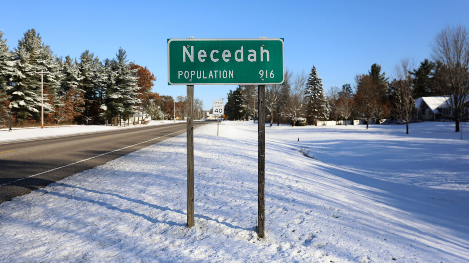 A municipality sign with the words Necedah and Population 916 stands on wood poles in a snow-covered lawn next to a road, with a speed limit sign, and snow-covered buildings and trees in the background under a clear sky. A municipality sign with the words Necedah and Population 916 stands on wood poles in a snow-covered lawn next to a road, with a speed limit sign, and snow-covered buildings and trees in the background under a clear sky.