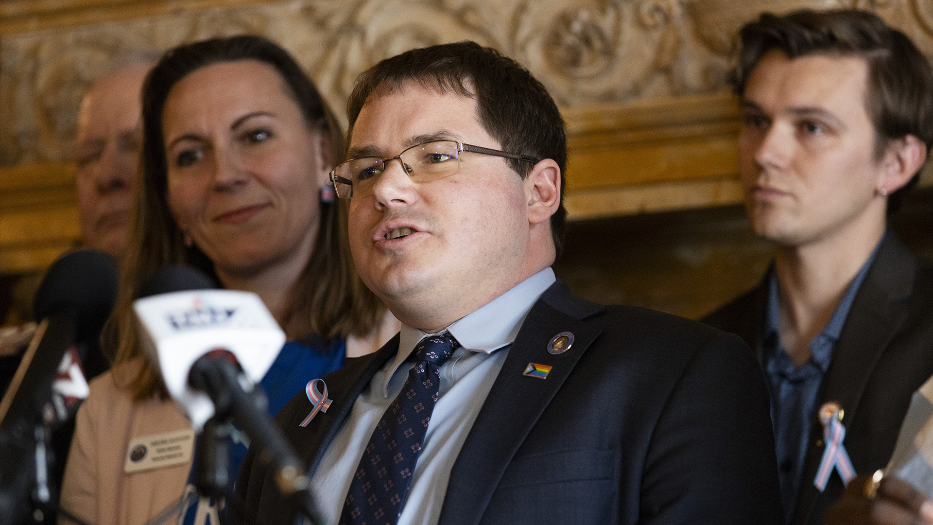 Mark Spreitzer stands and speaks into multiple microphones, with multiple out-of-focus people standing behind him in a room with a carved wood fireplace lintel.