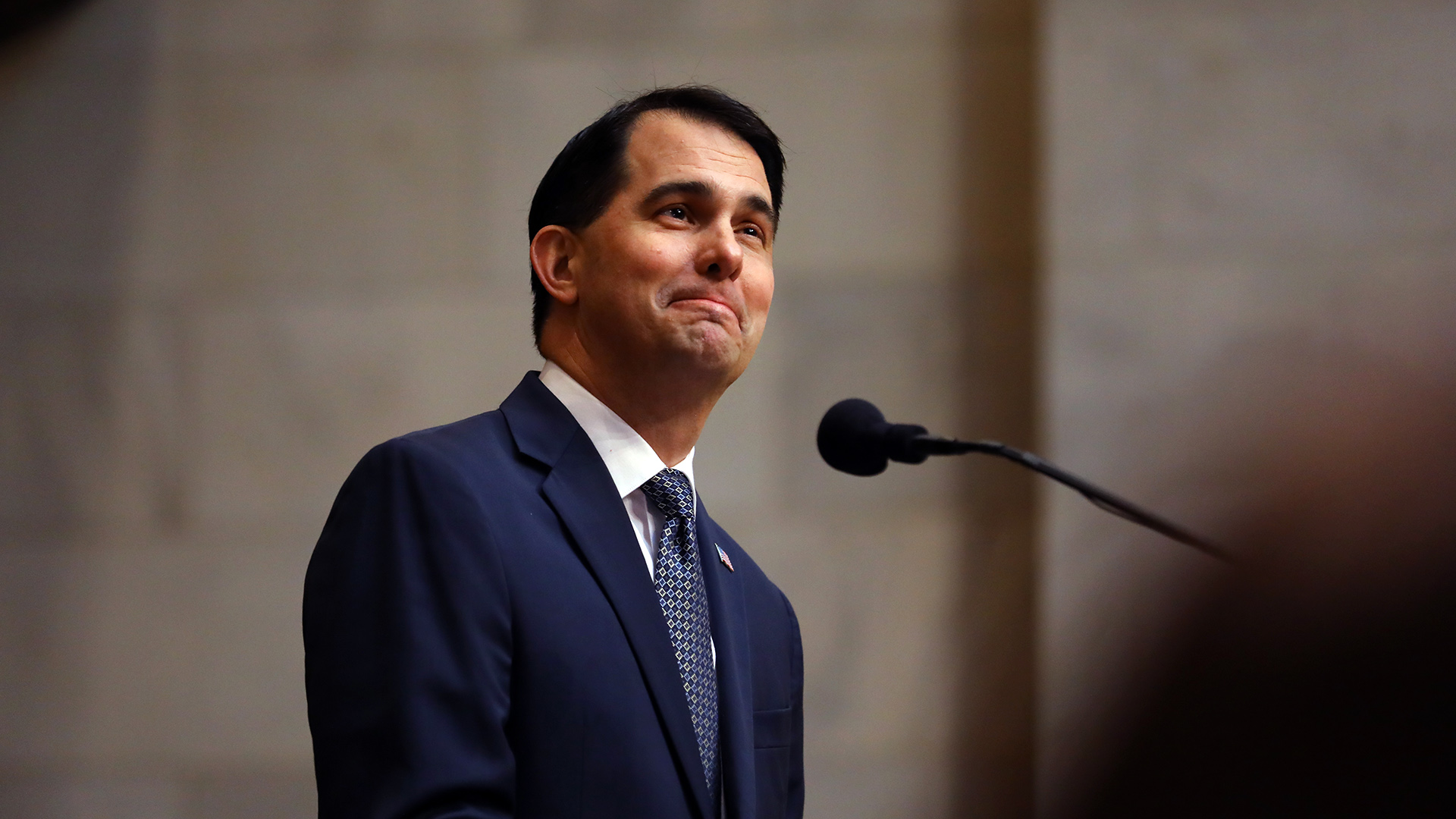 Scott Walker stands and speaks into a microphone on a mount, with an an out-of-focus head in the foreground and an out-of-focus marble masonry wall in the background.