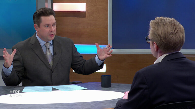 Two men in suits sit across from each other in a TV news studio, one gesturing broadly while speaking during a ‘Here & Now’ segment.