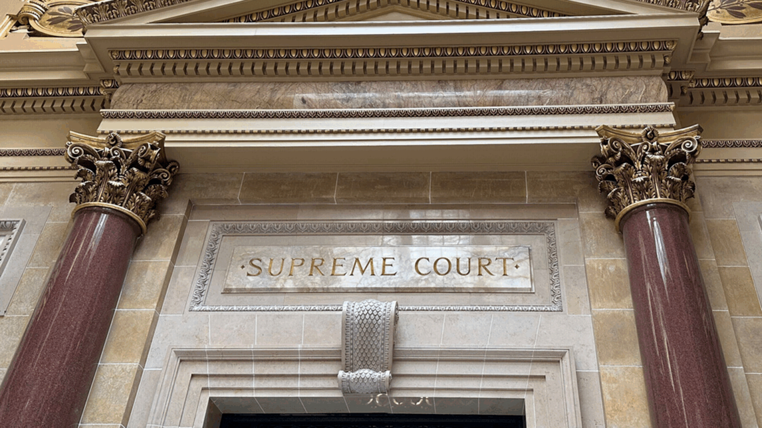 Ornate architectural entrance with red marble columns and gold-accented capitals, featuring the inscription SUPREME COURT. Ornate architectural entrance with red marble columns and gold-accented capitals, featuring the inscription SUPREME COURT.