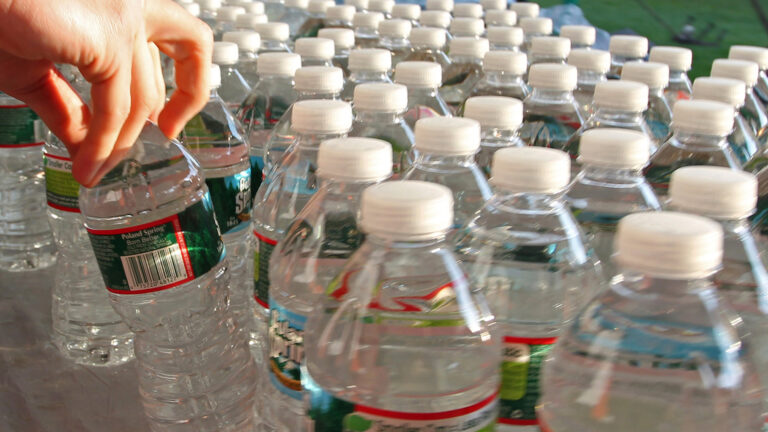 Hand reaching for a Poland Spring water bottle among rows of identical plastic bottles with white caps on a table.