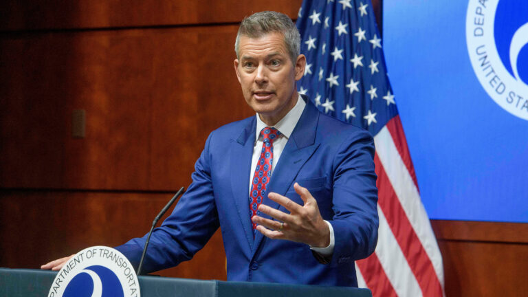 A man in a blue suit and patterned tie speaks at a Department of Transportation podium, gesturing with one hand; U.S. flag in background.