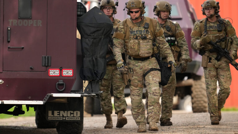 Heavily armed state troopers in camouflage tactical gear walk beside two armored vehicles.