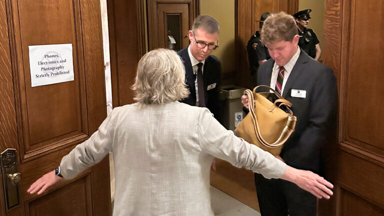 Security personnel conduct a bag check and pat-down at a wood-paneled doorway with a sign reading “Phones, Electronics and Photography Strictly Prohibited.”