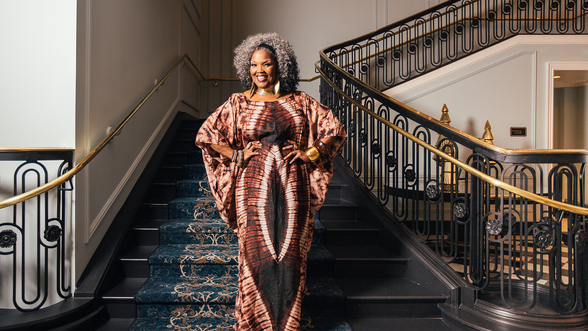 A woman with gray curly hair stands confidently on a grand staircase, wearing a patterned brown and black kaftan-style dress. She smiles brightly with her hands on her hips, accessorized with bold gold jewelry and bracelets. The staircase features ornate black and gold railings and a dark blue patterned carpet.