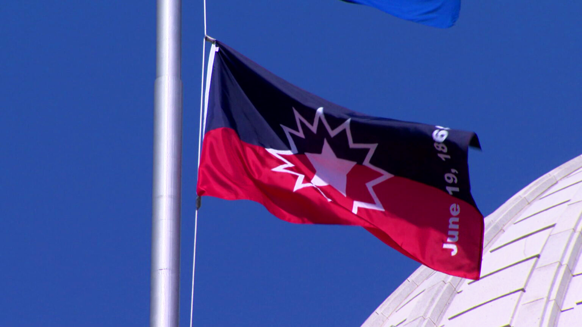 Red and black Juneteenth flag with a white starburst design and the text "June 19, 1865" flying on a flagpole against a blue sky.
