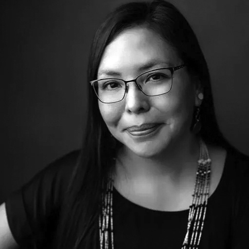 Jennifer Gauthier Black-and-white portrait of a woman with long hair, glasses, and beaded necklaces, smiling slightly against a dark background.