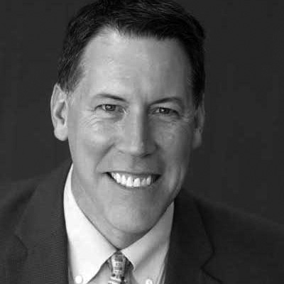 Terry Kaldhusdal Black-and-white headshot of a smiling man in a suit and tie against a dark background.