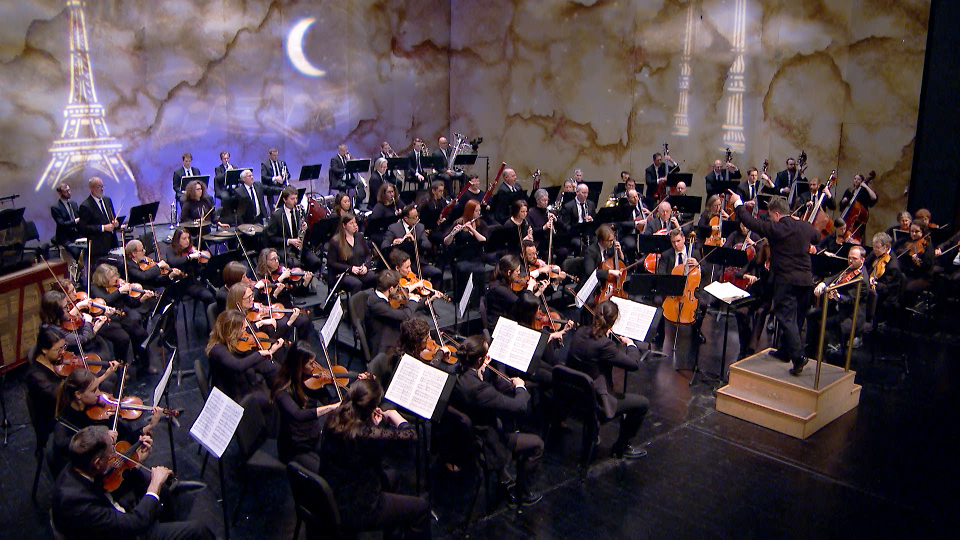 Alt text: A full symphony orchestra performs on stage, with musicians playing strings, woodwinds, brass, and percussion as a conductor leads from a podium. The backdrop features artistic projections of the Eiffel Tower, a streetlamp, and a crescent moon against a cloudy sky. 