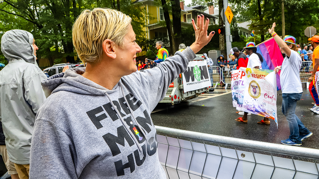 Person wearing a "Free Mom Hugs" hoodie waves at parade participants holding a colorful banner and rainbow flags on a rainy street.