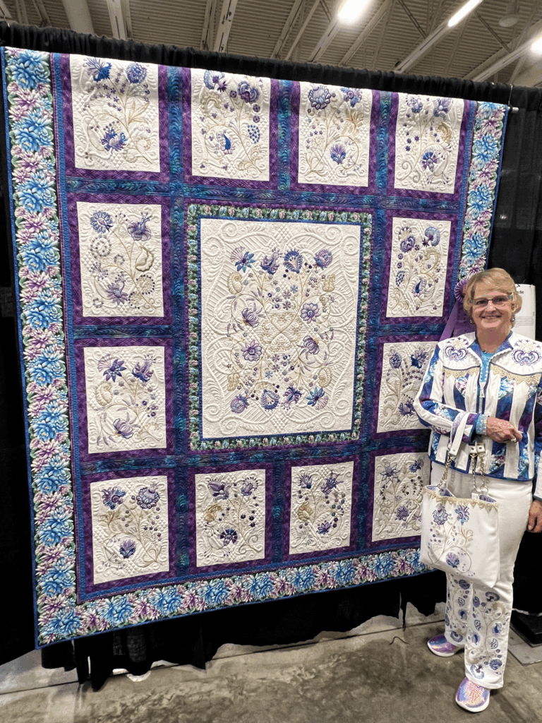 A woman dressed to match her intricate purple, blue, and white floral quilt stands proudly beside it at a quilt show.